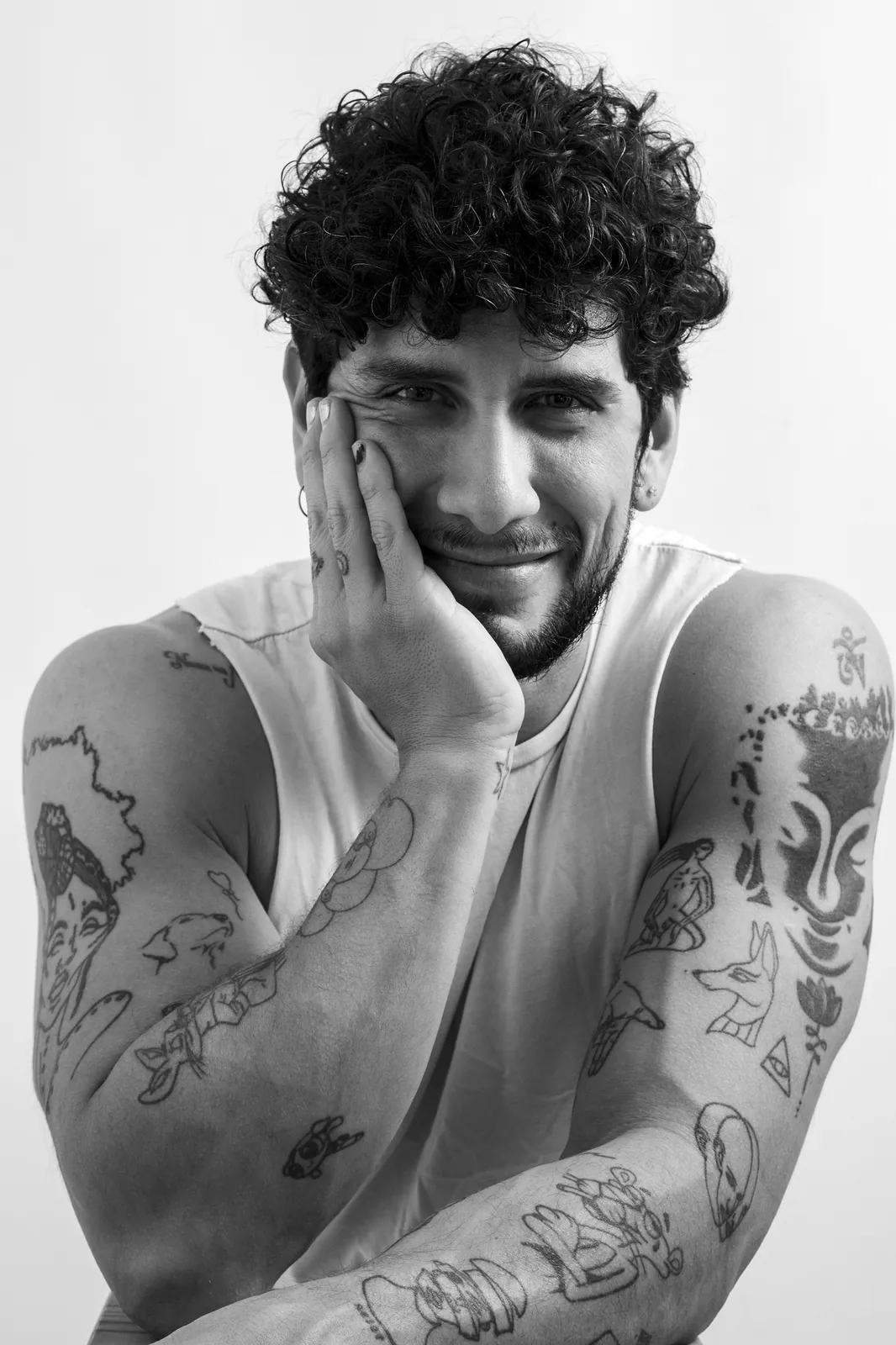 Retrato en blanco y negro de un hombre de pelo rizado y brazos tatuados, sonriendo con la mano en la mejilla